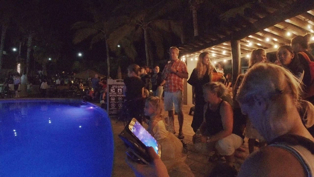 At 0:00 in Los Cabos, a blue-lit pool fills the left foreground while adults and children gather beside a warm-lit patio and a phone is held near the camera.