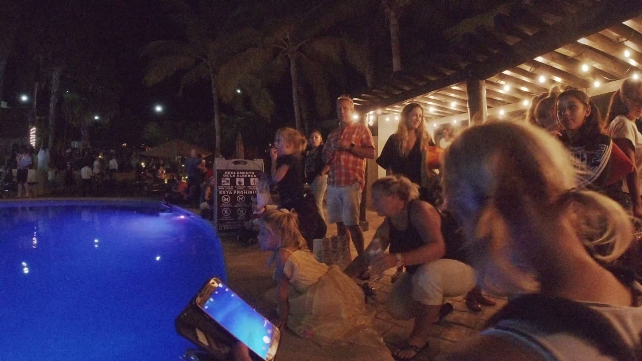 At 0:01 in Los Cabos, the pool remains on the left as a standing man, seated children, and several adults cluster near a signboard under warm patio lighting.