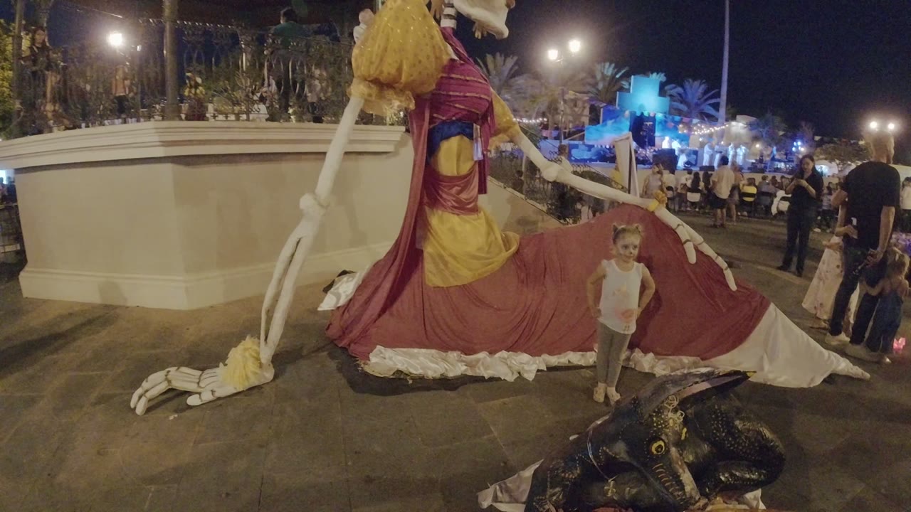 The child faces the camera in Los Cabos, Mexico with the skeleton sculpture's long white arm and red fabric behind, while a crowd stands across the plaza at night.