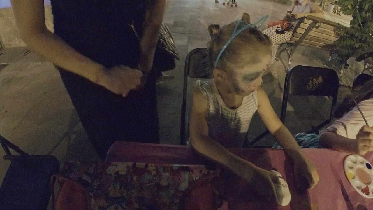 The girl with face paint reaches toward the table while another child paints beside a palette at the Los Cabos craft setup.