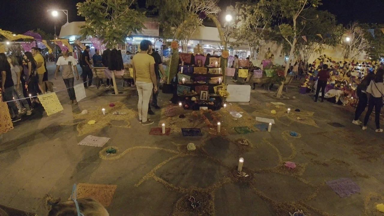 Los Cabos evening frame with a central memorial display, lit candles on the pavement, a man in a yellow shirt, and seated people gathered under trees and streetlights.