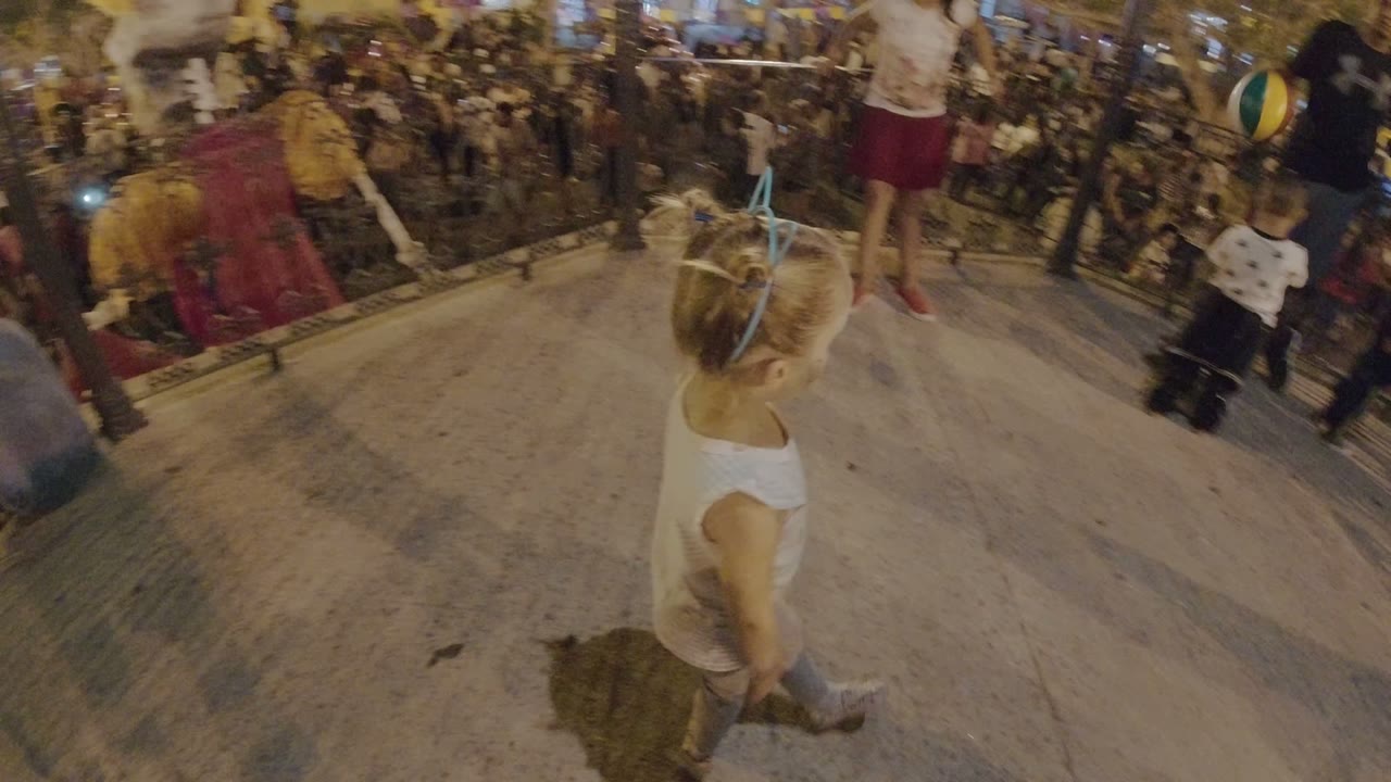 Young blonde girl seen from behind near a nighttime festival crowd.