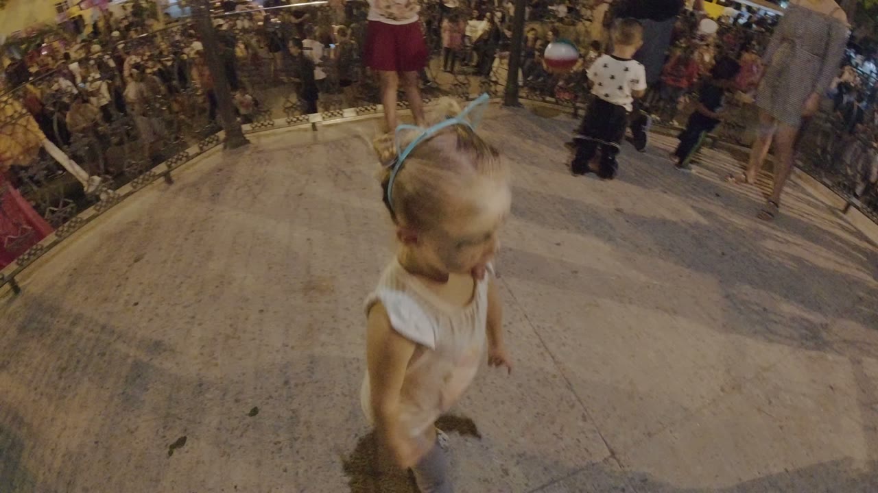 The blonde girl moves across the foreground while adults and other children stand near the railing at a busy Los Cabos nighttime festival.