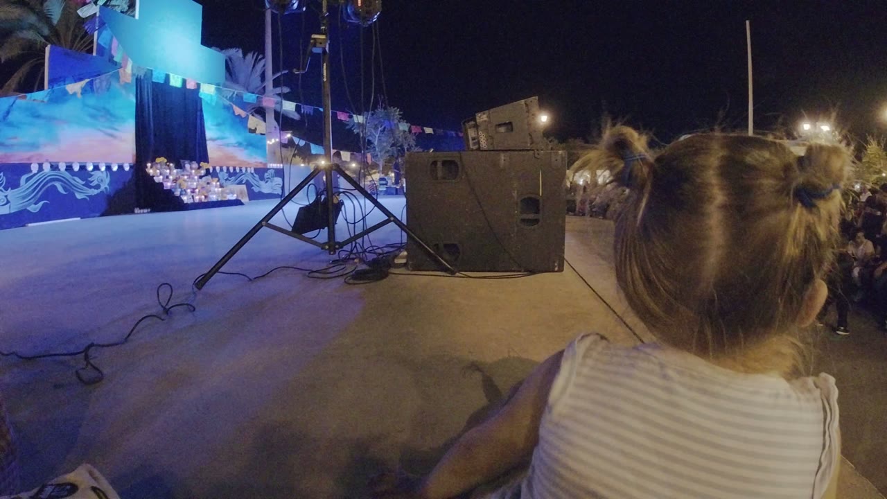 The child remains centered from behind at the edge of the Los Cabos stage, with blue lighting, a large speaker, and rows of spectators visible under night lights.