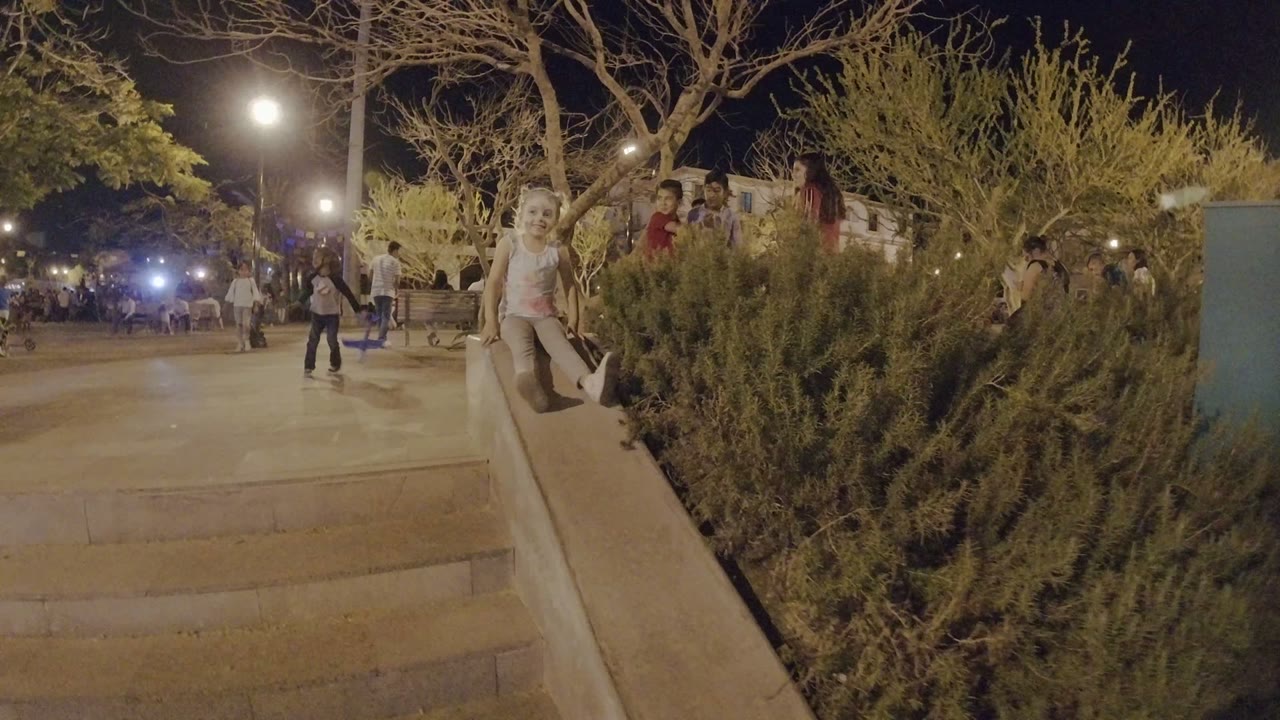 The girl begins sliding along the concrete handrail in Los Cabos, Mexico, with her legs forward and a shrub planter filling the right side of the frame.