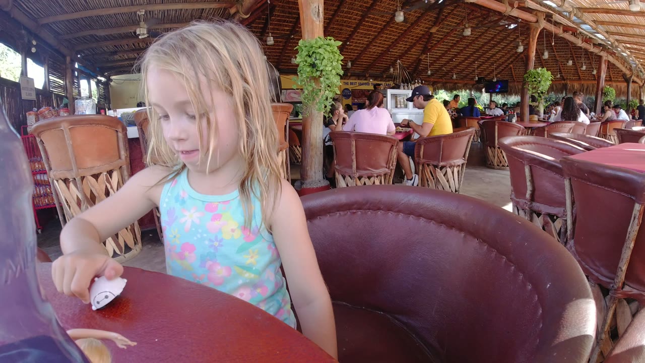 The child keeps her gaze down as she holds the small white object near the edge of the red table, with a curved leather chair and open-air restaurant seating in the background.