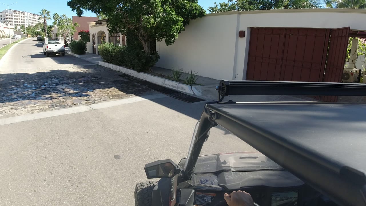 Vehicle roof and roll bar fill the lower frame as the view looks down a sunny residential street in Los Cabos, Mexico, with parked cars, a red gate, stucco walls, palms, and no visible people.