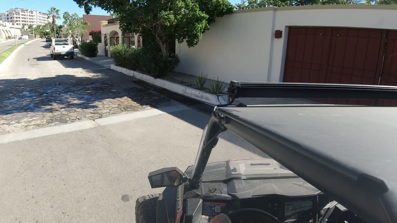 The moving vehicle passes a red gated wall in Los Cabos, Mexico, with a white car parked ahead, bright pavement, garden shrubs, and no visible people.