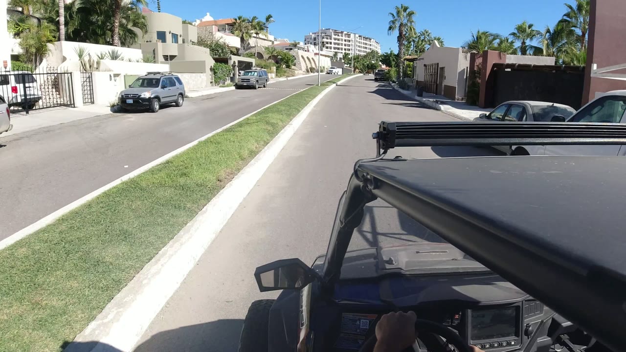 Residential street seen over a vehicle hood with homes, grass, parked cars, and palm trees.
