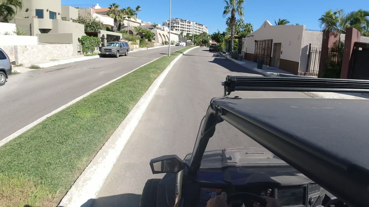 Second frame shows the Los Cabos street slightly farther along, with the vehicle hood in the foreground, a parked SUV near the curb, stucco homes, trimmed grass, palms, and no visible people.