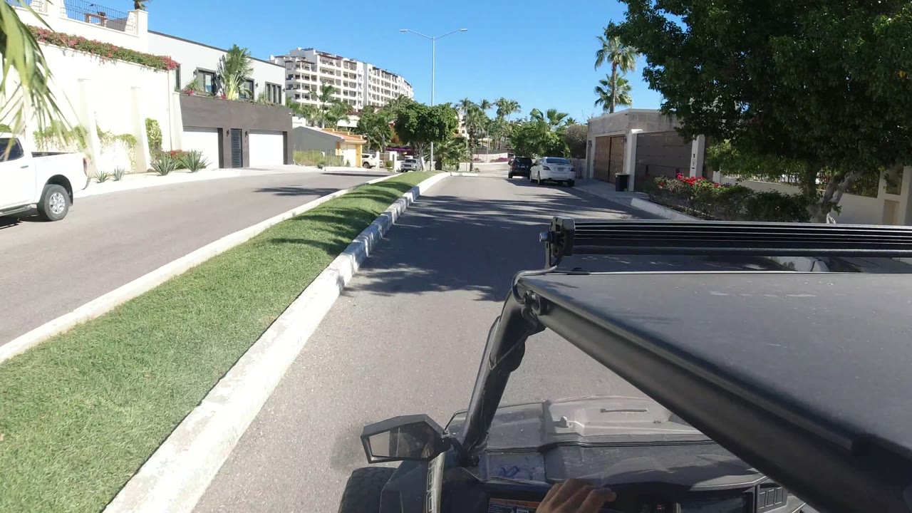Los Cabos street view with a moving-vehicle foreground, trimmed grass, modern white homes, palm trees, distant apartment buildings, and no visible people.