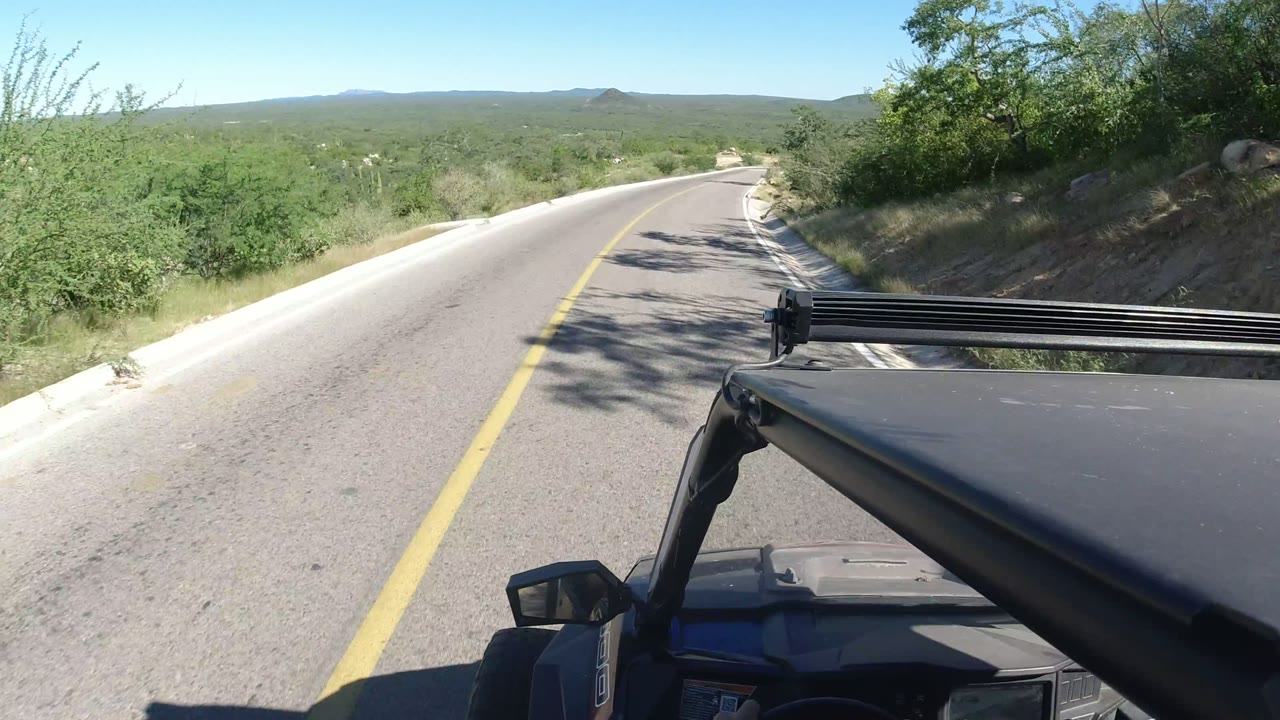 Los Cabos, Mexico road view one second in, with the yellow line leading around a bend, dry brush along the shoulder, and no visible people.