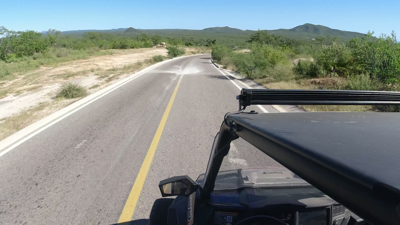 A Los Cabos road bends gently right ahead of the jeep, with the black light bar crossing the lower right foreground, pale dirt shoulders, green brush, distant mountains, and no visible people.