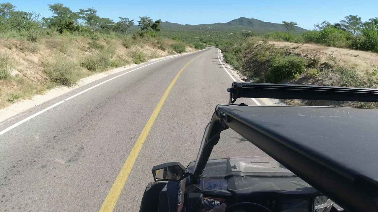 Los Cabos desert road viewed over a black off-road vehicle hood, with sandy shoulders, green scrub, low hills, blue sky, and no visible people.
