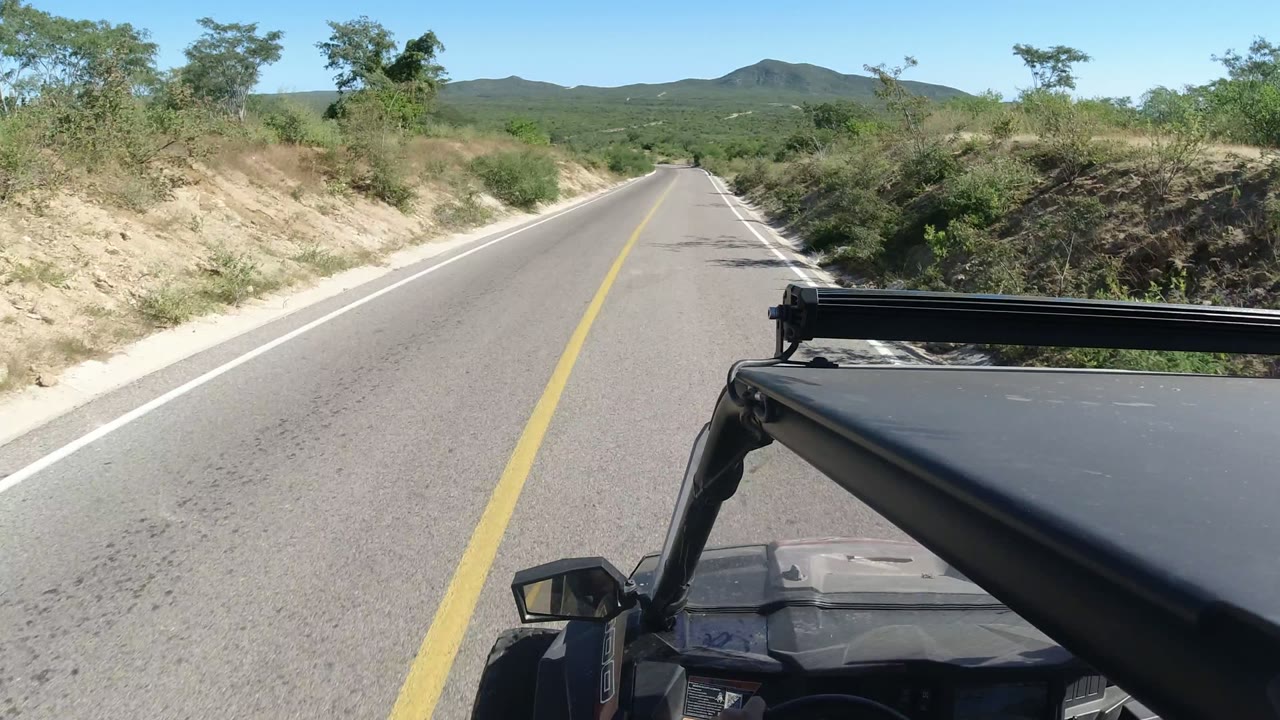 Forward view along a paved road in Los Cabos, with the vehicle front low in frame, scrub-covered banks on both sides, distant hills, and no visible people.