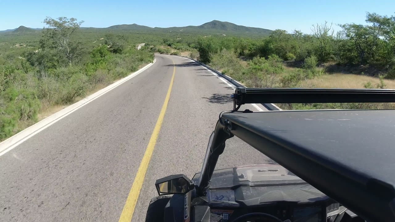 Forward view from a vehicle on a paved desert road in Los Cabos, Mexico, with a yellow center line, scrubby vegetation, distant hills, clear sky, and no visible people.