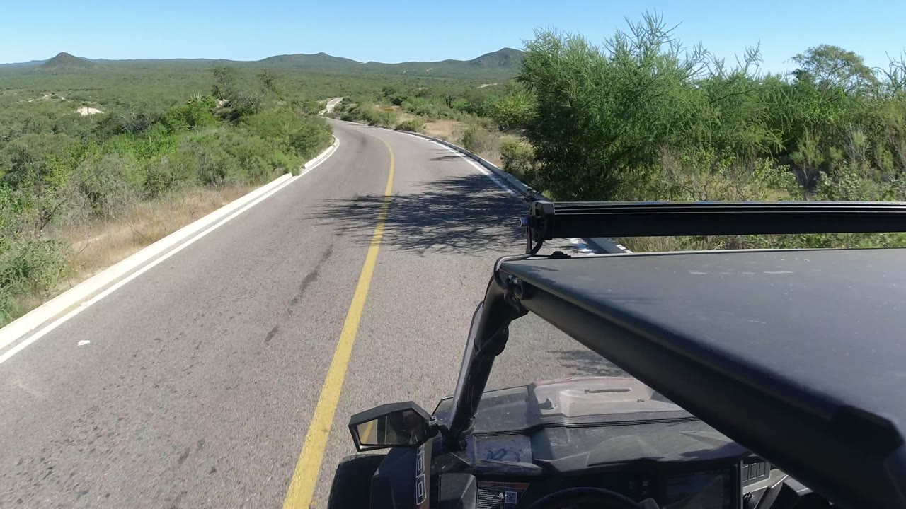 Vehicle-mounted view along a lightly curving paved road in Los Cabos, Mexico, with green desert shrubs close to the shoulder, low hills beyond, and no visible people.