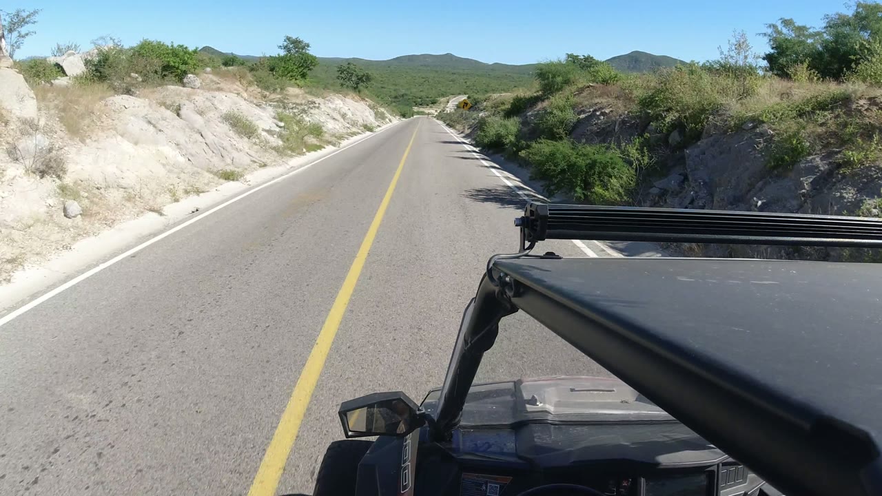 Vehicle-mounted view along a Los Cabos road as the asphalt runs between pale sandy roadside cuts, green shrubs, distant hills, and no visible people.