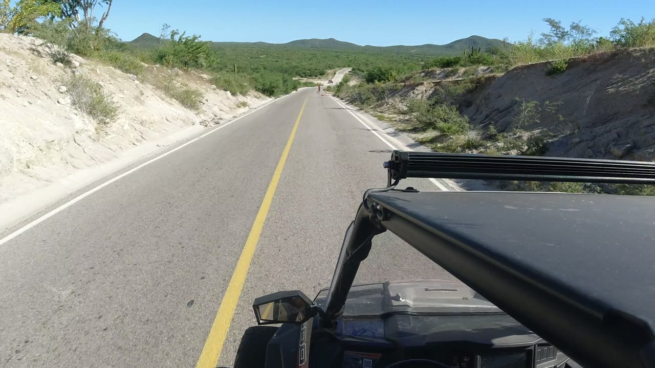 At 0:00, a Los Cabos road stretches ahead between pale roadside banks and scrub vegetation, with the vehicle roof and side mirror visible in the foreground and no visible people.