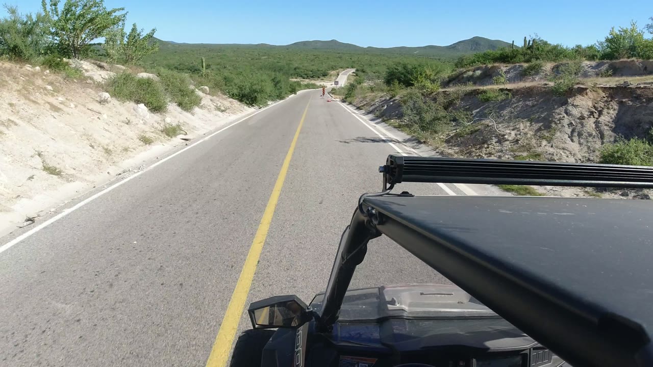 At 0:01, the road continues through Los Cabos scrubland, framed by the vehicle canopy in the lower foreground, pale shoulder cuts, distant hills, and no visible people.