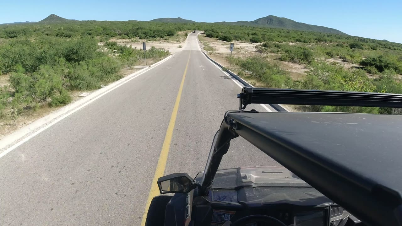 The SUV point of view continues along the Los Cabos road, passing green brush and roadside posts under bright midday light with no visible people.