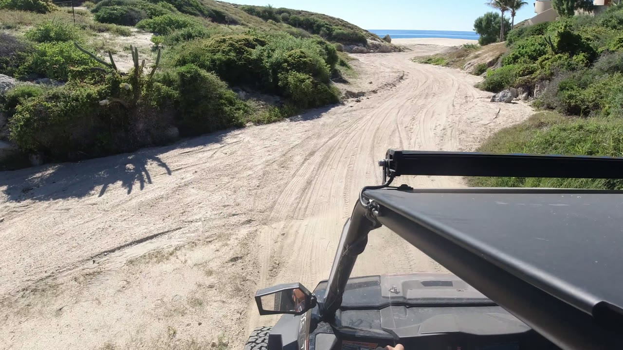 The jeep continues along the pale sand road in Los Cabos, with the vehicle roof bar low in frame and the ocean line centered beyond the dunes.