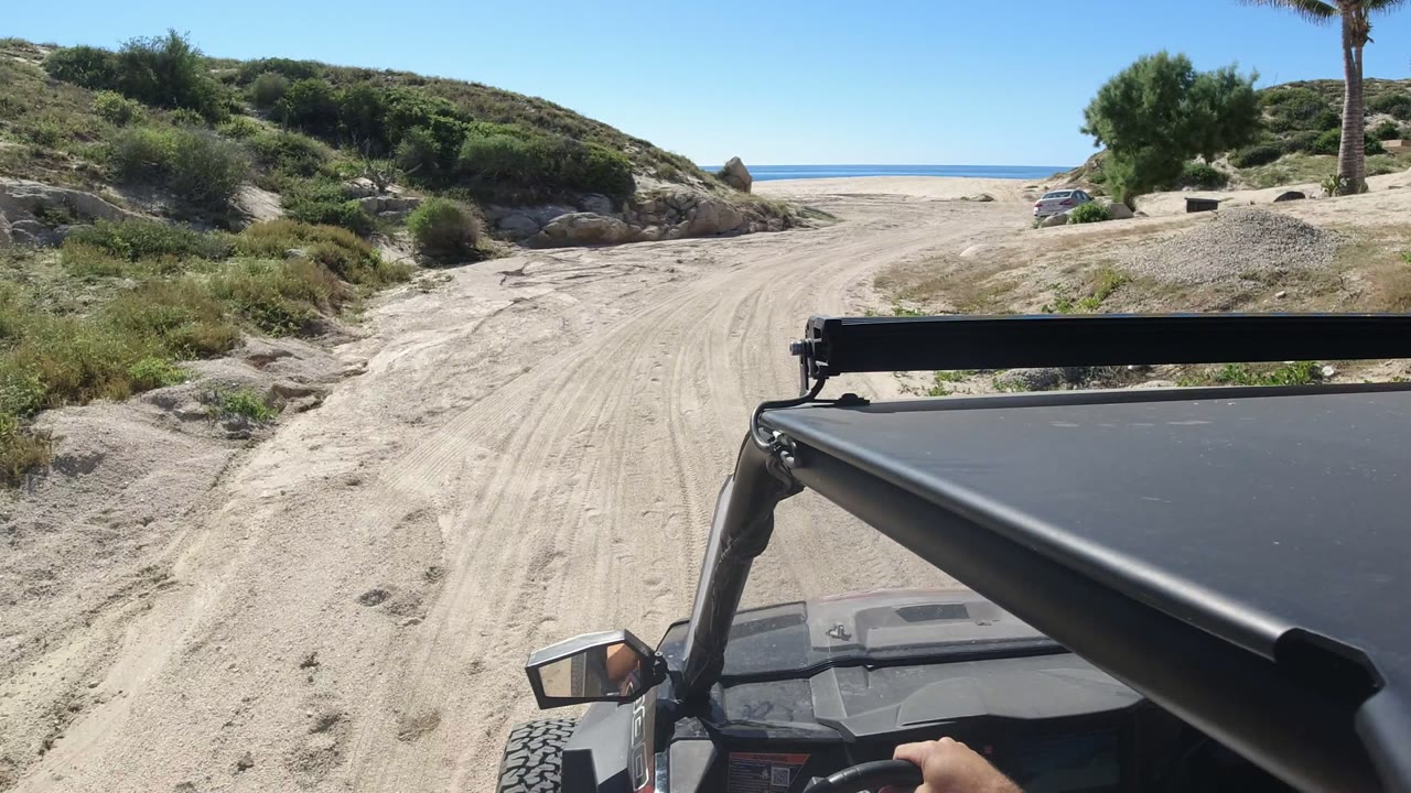 Los Cabos sandy beach track seen from an off-road vehicle, with the black canopy and mirror in front, low brushy hills on both sides, ocean in the distance, and no visible people.