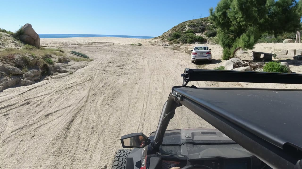 Black off-road vehicle approaching a sandy beach with a white car near dunes.