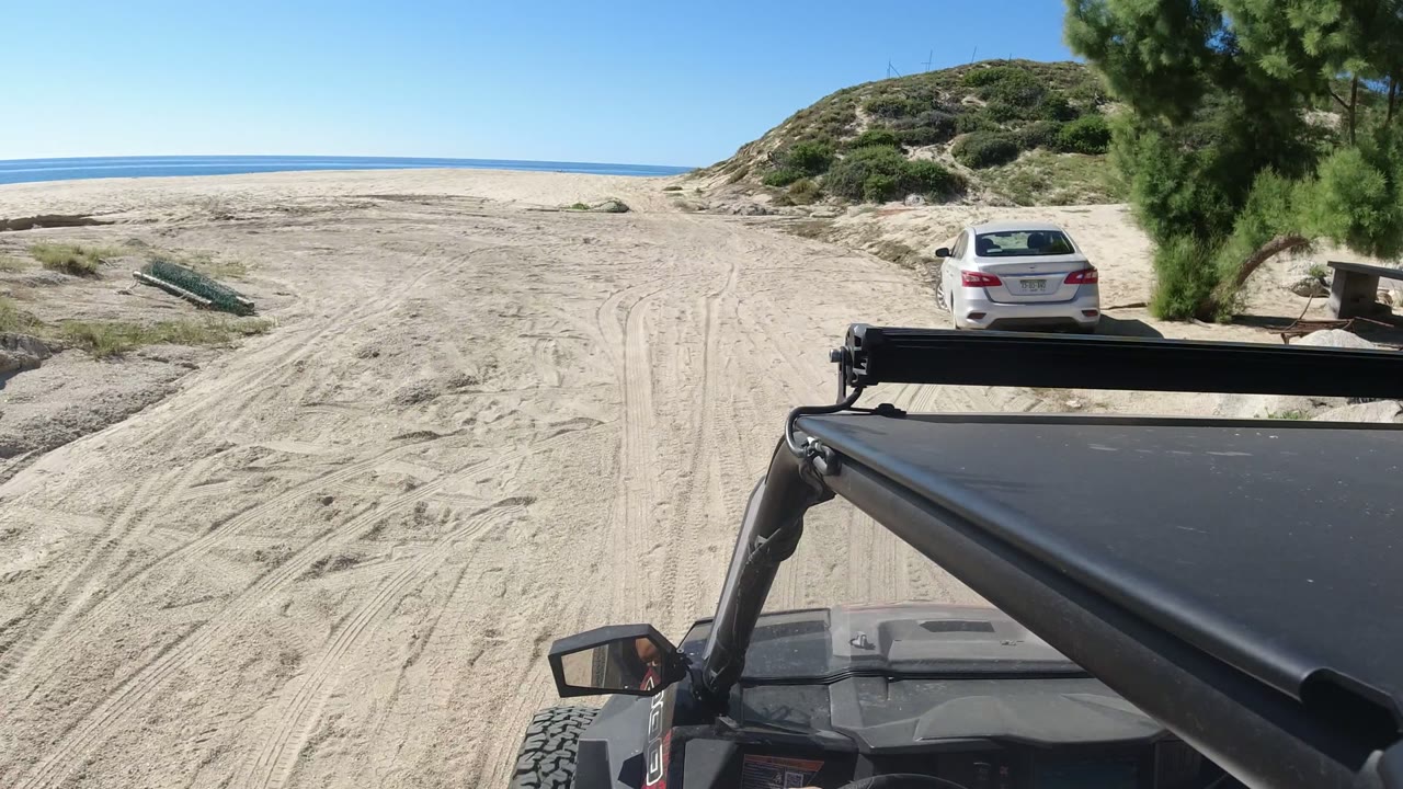 Vehicle POV in Los Cabos, Mexico, showing the black vehicle foreground, tire-marked sand ahead, a white car near a tree to the right, dunes, ocean, and no visible faces.