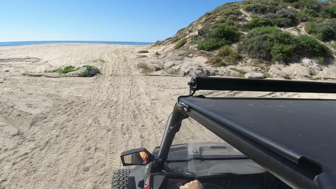 The Jeep foreground continues along a Los Cabos beach track, with pale sand ahead, scrub-covered dunes to the right, and open ocean visible beyond.