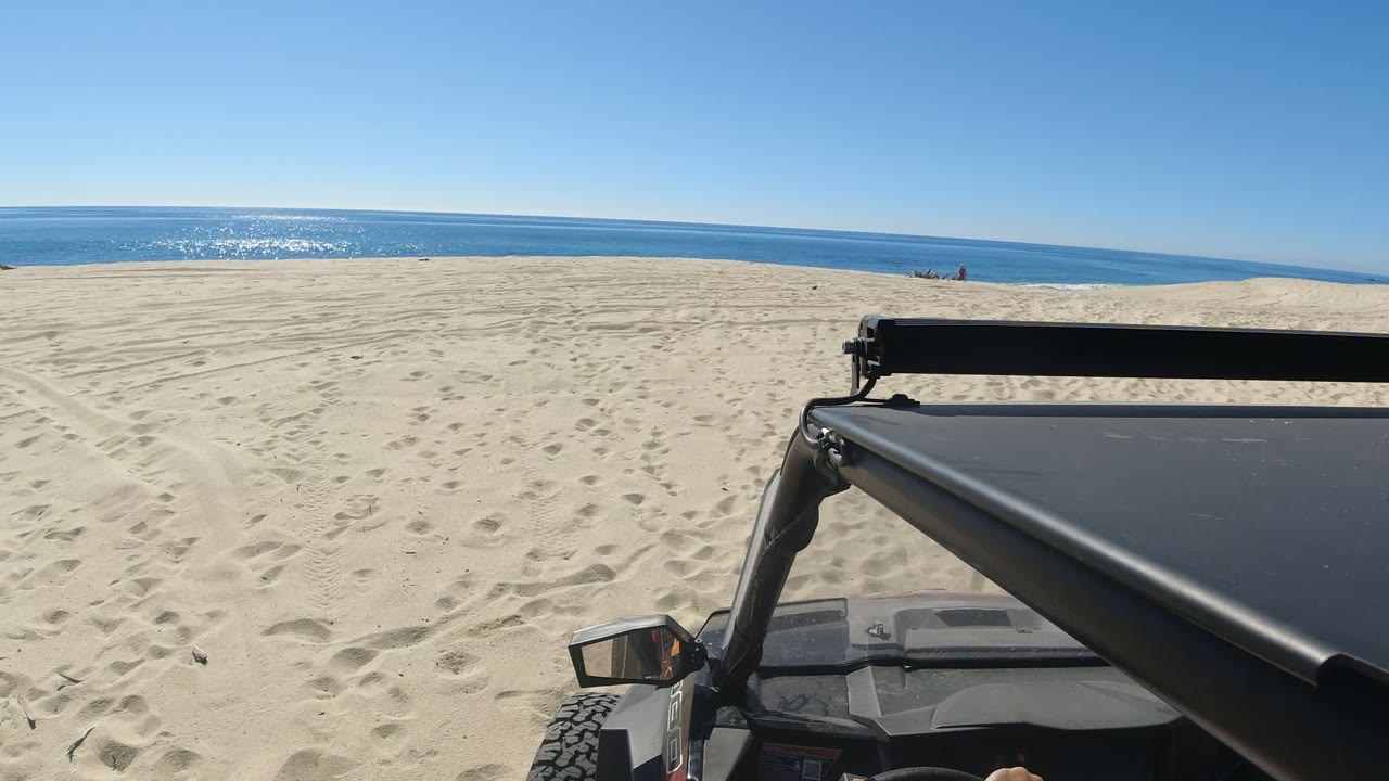 The second frame keeps the Los Cabos beach horizon centered while the vehicle roof and roll bar occupy the lower right foreground and faint distant figures sit near the waterline.