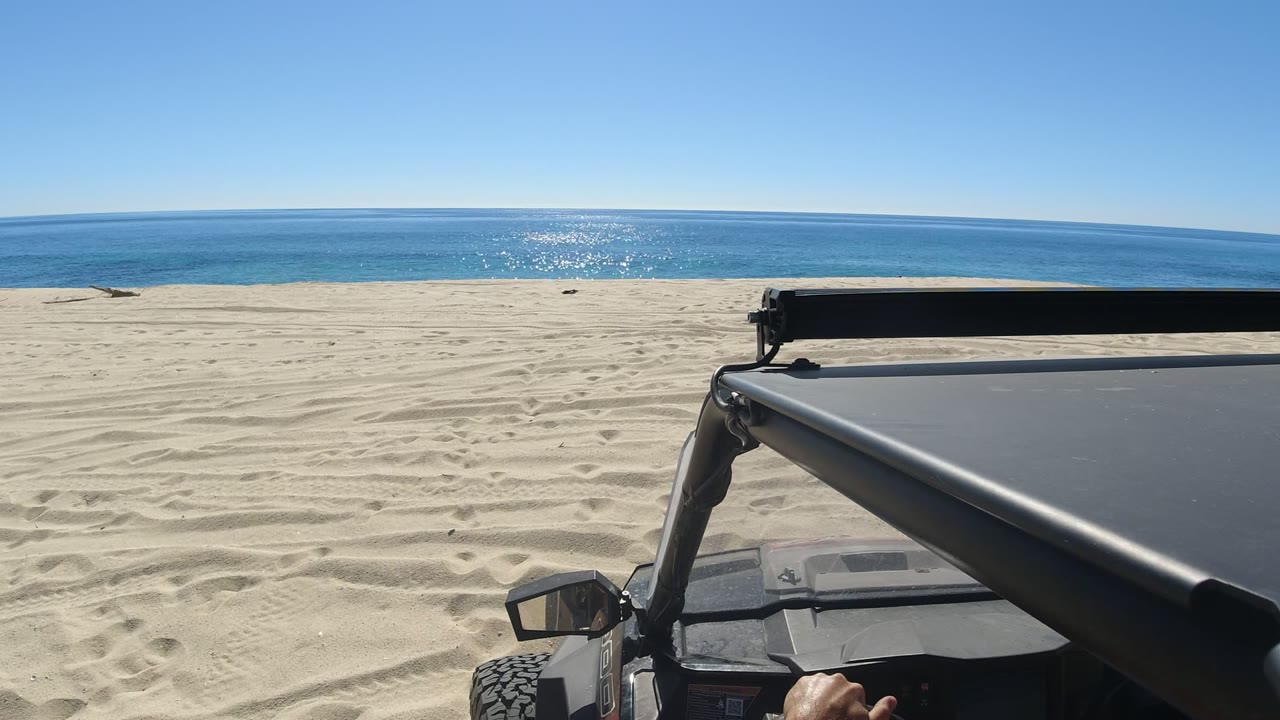 Los Cabos beach view at one second with the vehicle roof occupying the lower right, textured sand across the foreground, bright blue water on the horizon, and a small hand visible near the dashboard.