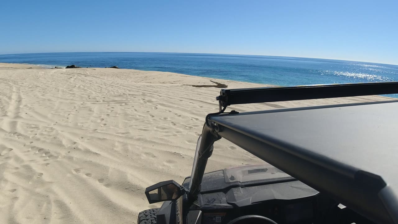 A sandy track runs toward the blue ocean in Los Cabos, Mexico, while the black vehicle hood and roof bar occupy the lower right foreground with no visible people.