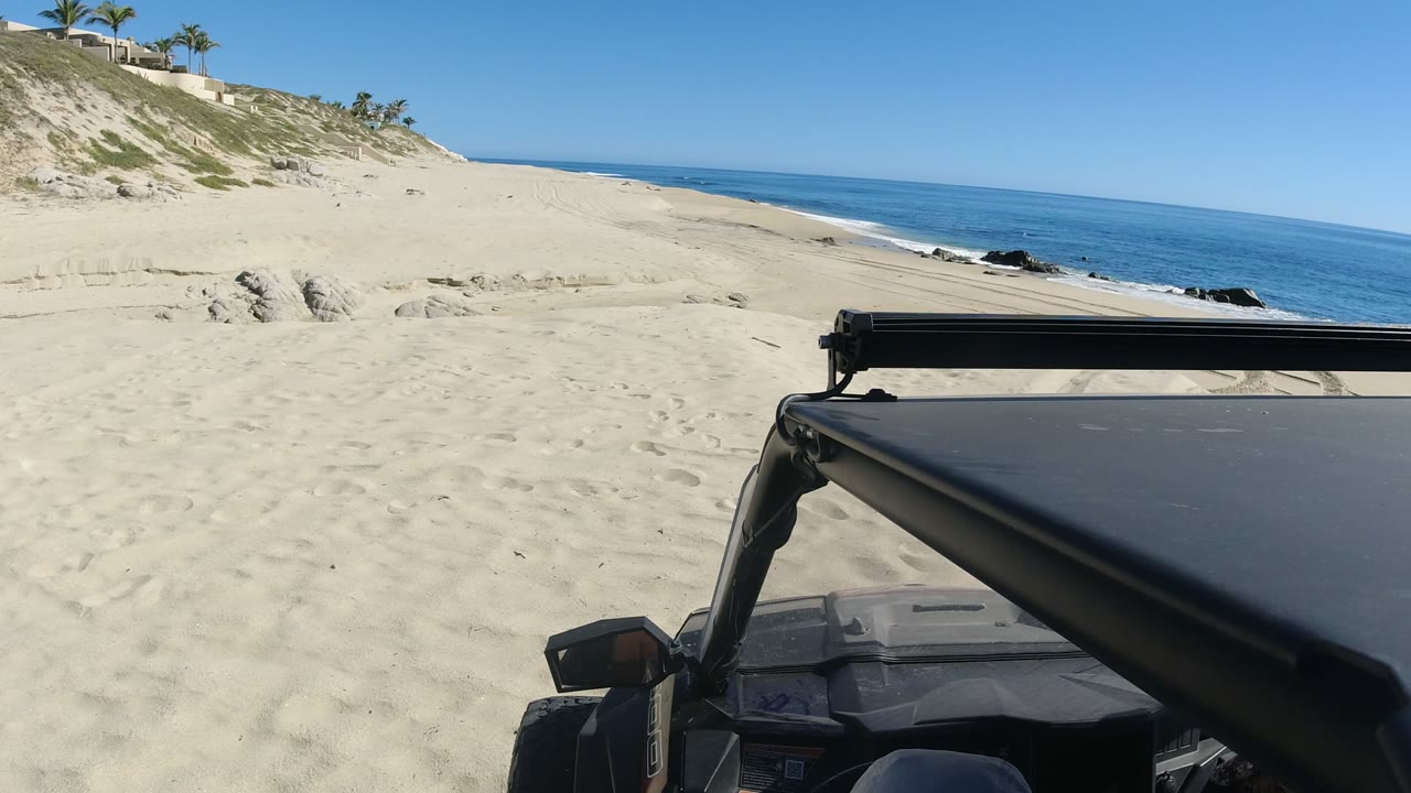 At 0:01, the Jeep remains in the lower right while the sandy Los Cabos shoreline stretches ahead, with small surf lines, dark rocks near the water, hillside palms, distant homes, and no visible people.