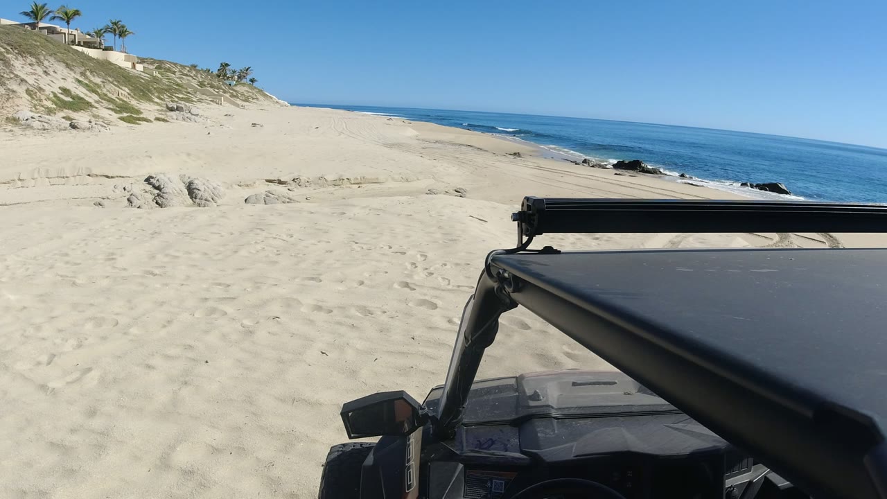 Sandy beach and ocean seen past the front of a dark vehicle.