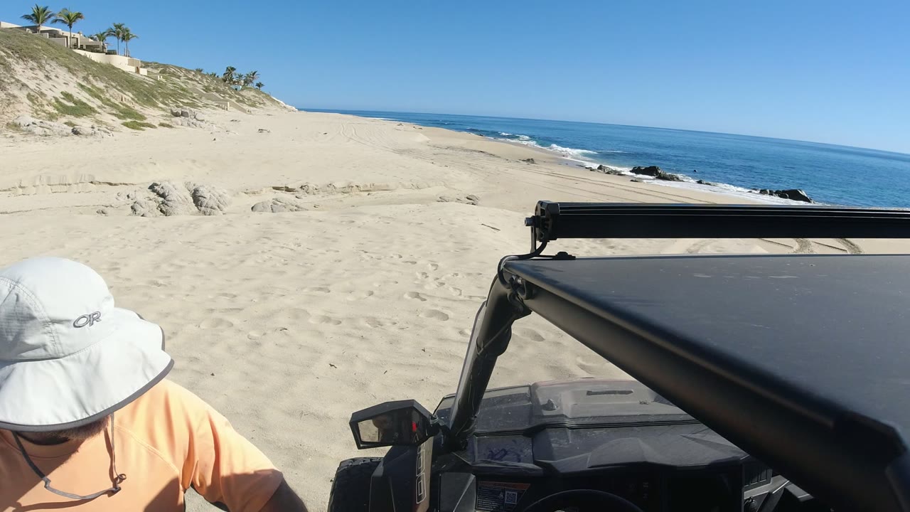 The person leans down at the edge of frame beside the dark vehicle, with Los Cabos sand, low dunes, ocean water, and a bright horizon visible beyond.