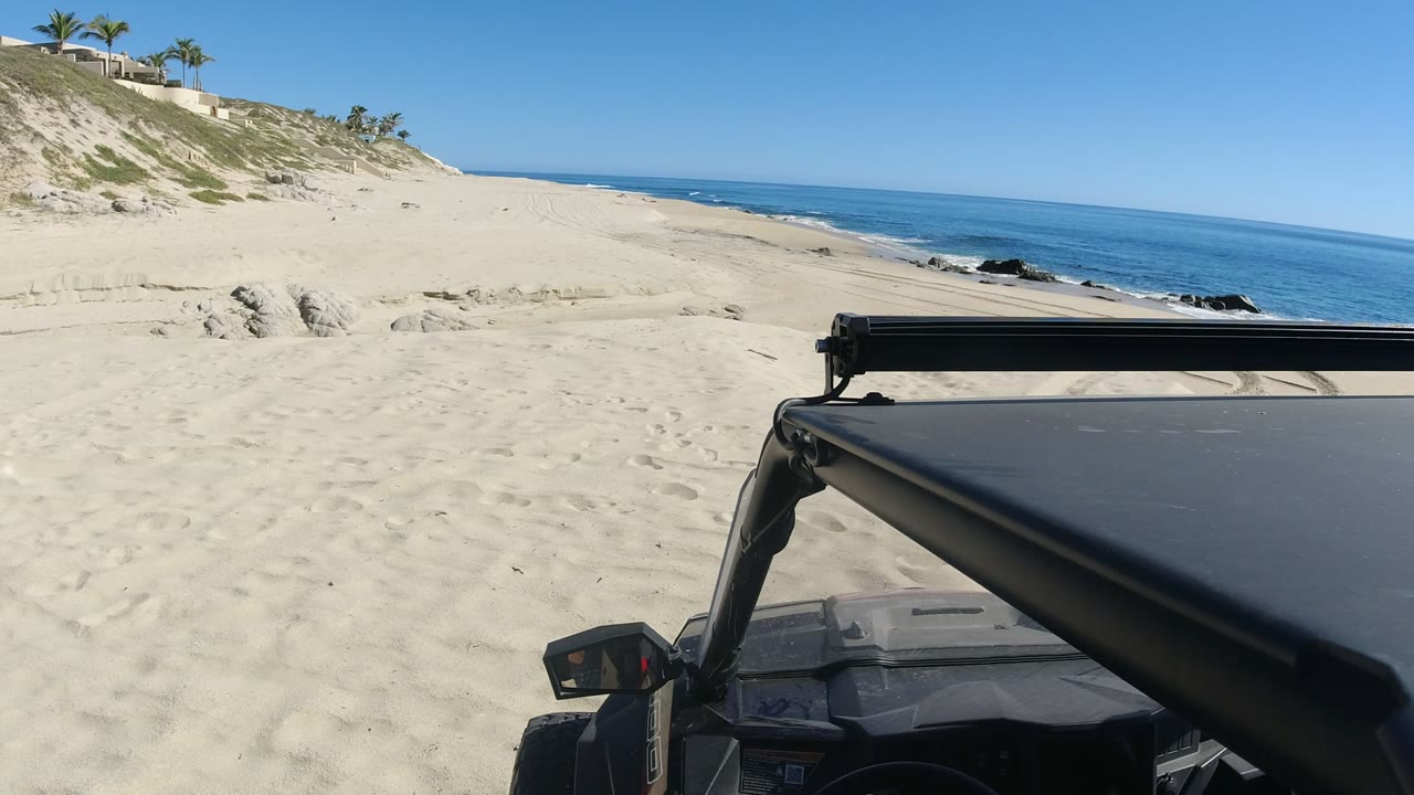 Black vehicle roof beside a sandy Los Cabos beach and blue sea.
