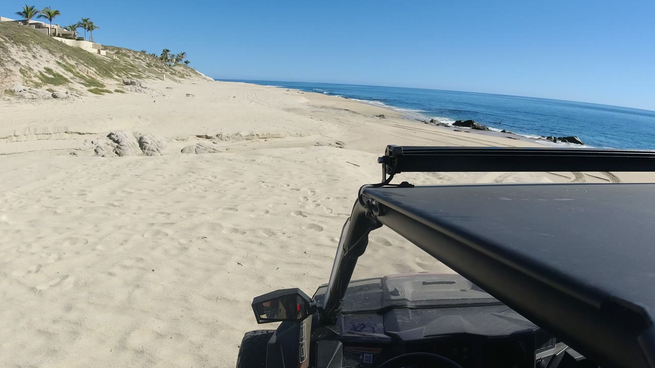 At 0:01, the black vehicle remains in the right foreground as pale beach sand, a sloped dune with buildings, dark rocks near the water, and the Los Cabos sea fill the frame with no visible people.