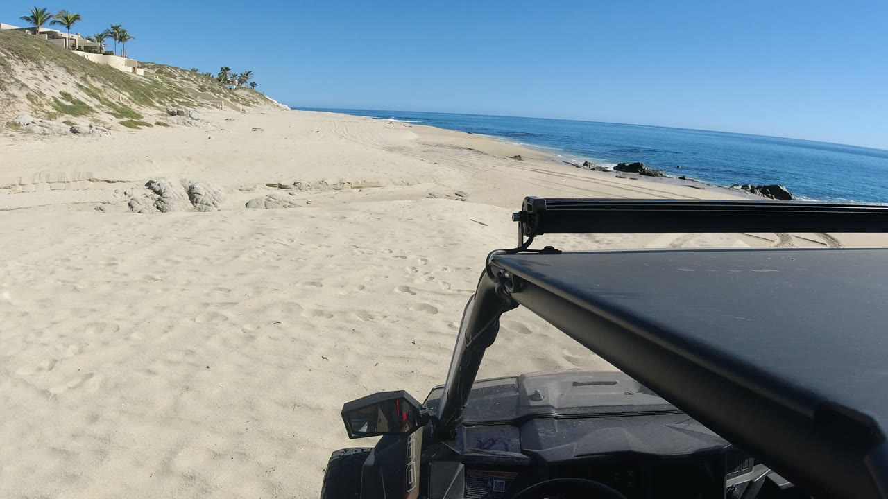 At 0:00 in Los Cabos, a vehicle roof and roll bar fill the lower right foreground while pale sand, dunes, black shoreline rocks, and blue ocean sit beyond, with no visible people.