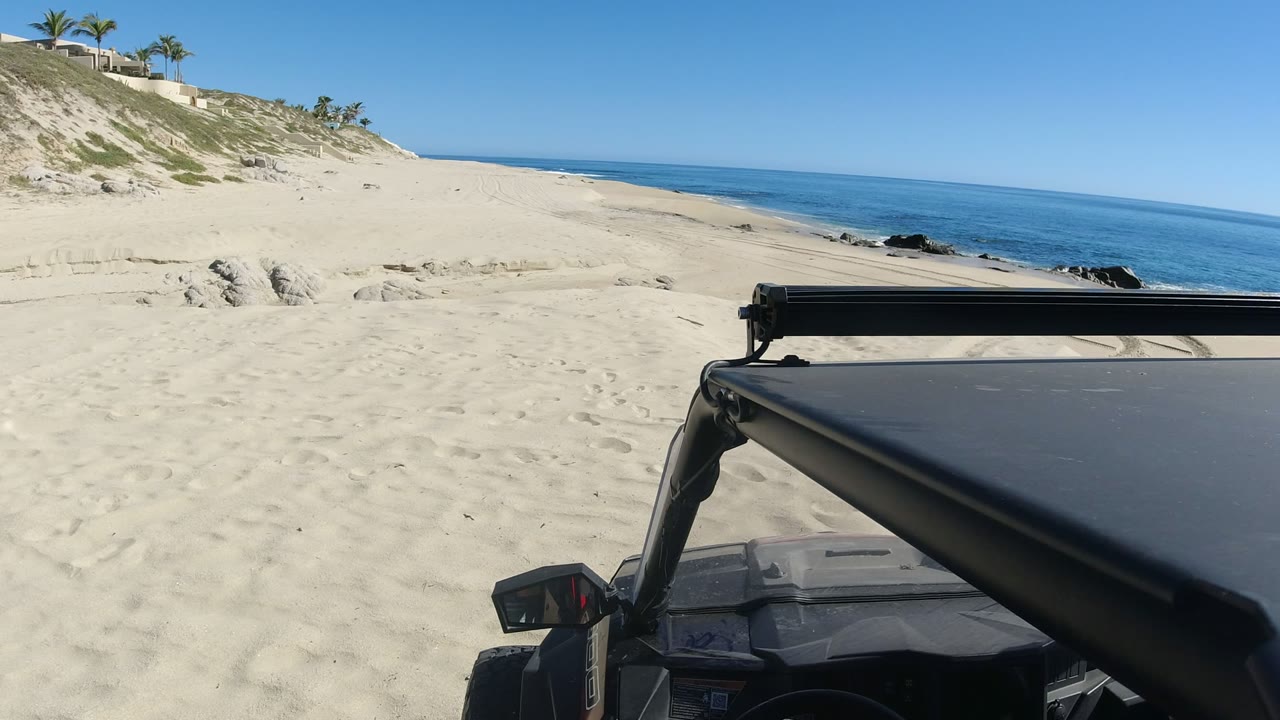 At 0:01 in Los Cabos, the vehicle roof and frame remain at lower right with broad empty sand, dune fencing, palms, distant buildings, ocean, and black rocks visible, with no visible people.