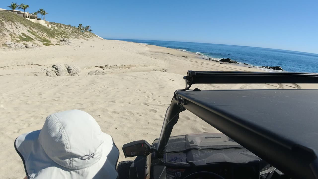 At 0:00 in Los Cabos, Mexico, a sandy beach and blue ocean appear beyond a black off-road vehicle roof, with two hat-covered people partly entering the lower frame.