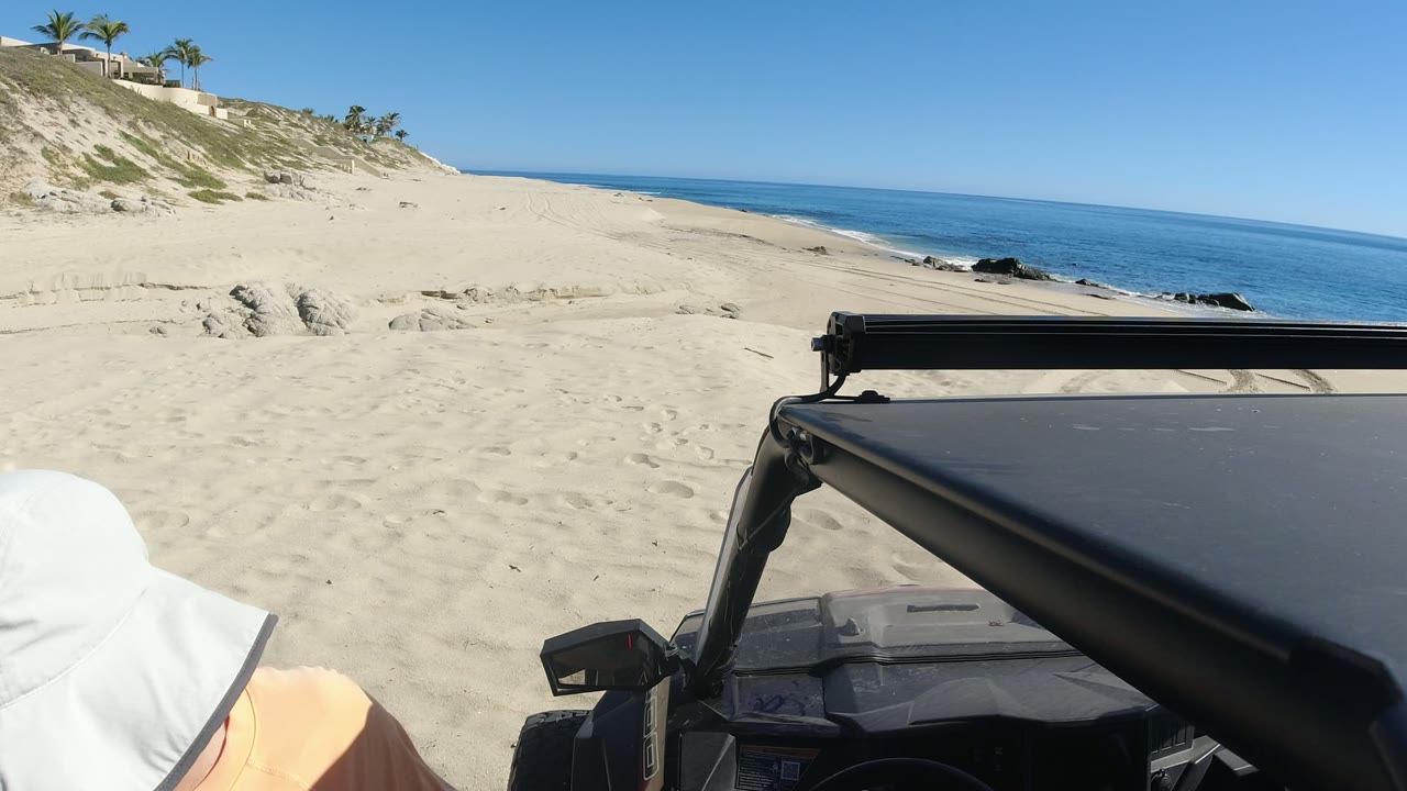 At 0:01 in Los Cabos, Mexico, the frame centers on pale sand, a blue ocean horizon, and the black roof and roll bar of an off-road vehicle, with no visible people.