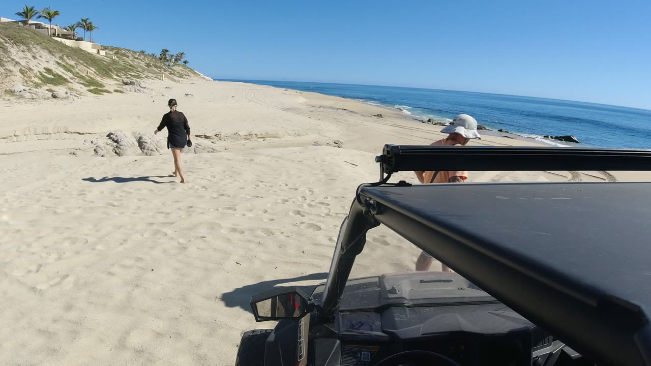 In Los Cabos, Mexico, a person walks away across pale sand while a black off-road vehicle roof fills the right foreground and the blue ocean sits beyond the beach.