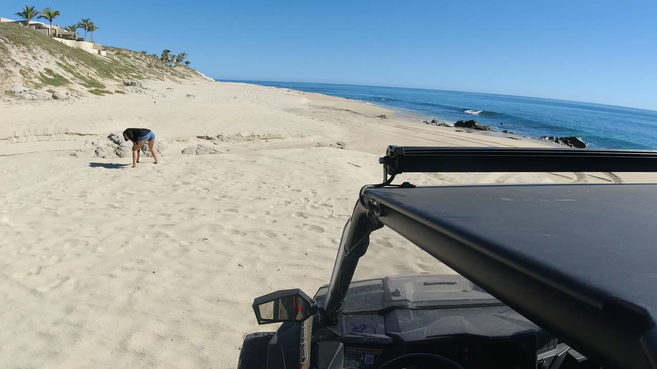 Woman bending over on a sandy beach beside ocean waves and a foreground vehicle.