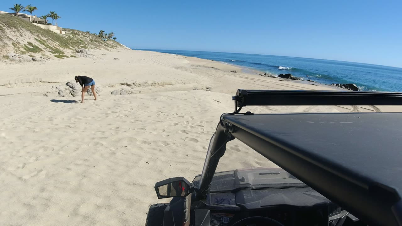 One second into the Los Cabos beach clip, the woman remains bent near a disturbed patch of sand as the shoreline and vehicle stay in the same composition.