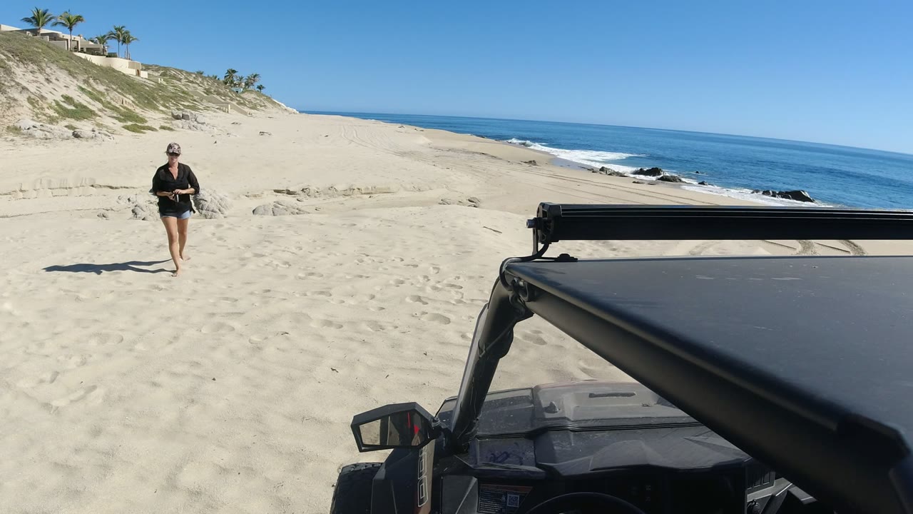 Woman walking on a sandy beach near an off-road vehicle and ocean shoreline.