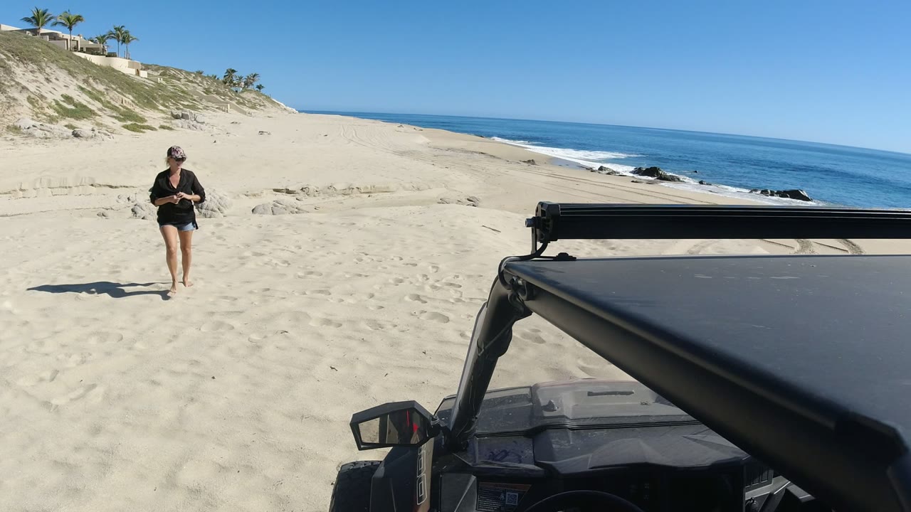 The woman moves closer across the Los Cabos beach, holding items in her hands while the parked dark vehicle stays in the foreground and small waves break along the shore.