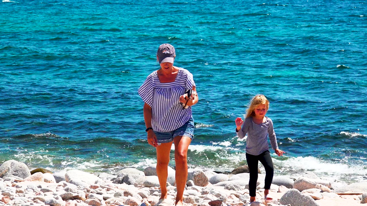At 0:00, a woman in a striped shirt and cap walks near a blonde child on the rocky shore in El Sargento La Ventana, Mexico, with blue water behind them.
