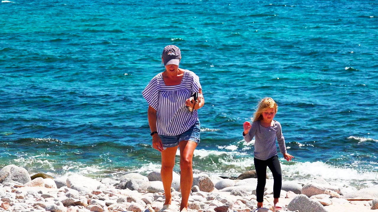 Woman and child continue walking across stones near the surf.