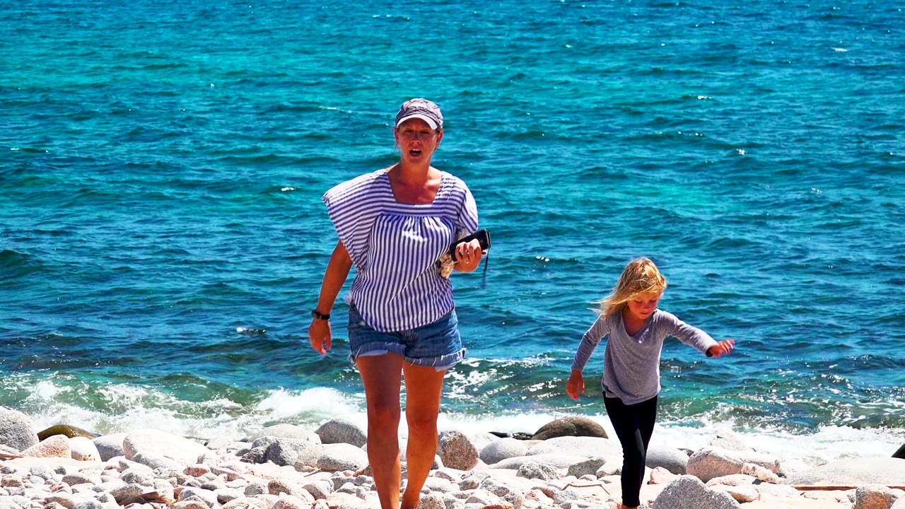At 0:02, the woman and child move toward the camera across the rock beach, with the woman lifting one foot and the child balancing near the edge of the water in El Sargento La Ventana, Mexico.